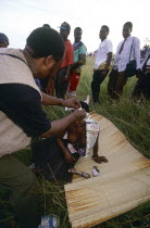Zulu man pinning money to a girls head whilst she sits on a mat in a field at a coming of age ceremony.