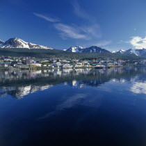 The town of Bahia Ushuaia reflected in still water with distant snow capped mountains in a typical Patagonian landscape
