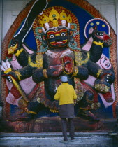 Man in front of temple carving of Black Bhairab in Durbar Square depicting Shiva in his most fearsome form with six arms  a head dress of skulls and carrying weapons.