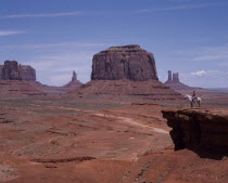 Mittens desert rock features. Isolated blocks and pinnacles and flat valley floor with road running through. Rider on horse admires view from rocky ledge.
