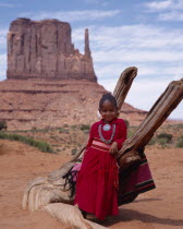 Navejo girl leaning against dead tree wearing a red dress and turquoise necklace and broach with the Mittens rock formation in the background