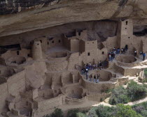 Cliff Palace the preserved ruins and cave houses beneath rock face. Visitors walk through site of walls and towers with circular foundations leading to deeper caves.