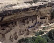 Cliff Palace the preserved ruins and cave houses beneath rock face. Visitors walk through site of walls and towers with circular foundations leading to deeper caves.