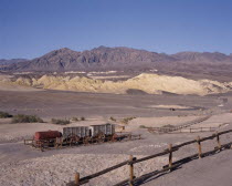 Historic Harmony Borax Works. Wooden containers and steel cylinder on trailers in fenced area in desert landscape with hills in the background