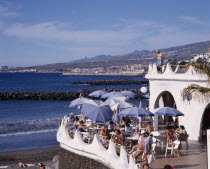 Playa de las Americas. Terraced bar overlooking the beach with customers sitting at table and a man standing on a balcony above