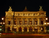 Opera frontage floodlit at night against black sky with light trails from passing traffic in the foreground.