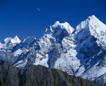 Himalayan mountain peaks with moon above just visible in deep blue sky.