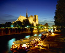Notre Dame floodlit at night with restaurant and houseboat lit up on the River Seine in foreground.