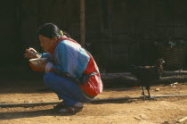 Lisu woman eating whilst squatting outside her home with a chicken behind her