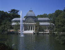 Retiro Park. The Crystal Palace surrounded by trees seen from across pond with fountain.