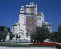Plaza Espana. Stone obelisk with statue of Miguel de Cervantes surrounded by trees and flower beds with tall office building behind.