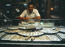 Jeweller behind display of gold items in glass topped cabinet.
