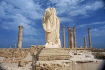 Headless and armless classical statue on raised plinth with standing columns behind.