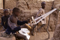 Child weaving on hand loom in Dogon village with other children behind.