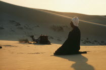 Muslim man praying in the desert with camel and sand dunes behind. Moslem