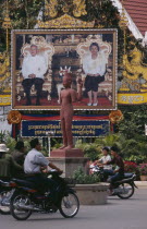Poster of Sihanouk and his wife by the side of a roundabout with motorbikes going around an armless statueAsian Cambodian Kampuchea Southeast Asia Kamphuchea