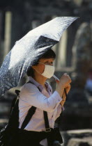 Cambodia, Siem Reap Province, Angkor Thom, Japanese tourist at Bayon Temple, wearing face mask and holding umbrella as sun shade.