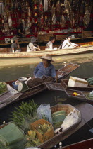 Damnoen Saduak Floating Market with an old female fruit vendor in her canoe and tourists in a canoe behind in front of a tourist goods stall