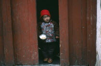 Child holding bowl of rice and chopsticks looking out from narrow doorway.