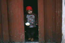 Child holding bowl of rice and chopsticks looking out from narrow doorway.