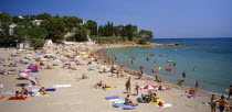 Curved  sandy beach with people sunbathing and in the sea.  White painted houses amongst trees on hillside beyond.  Costa Brava