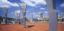 Port Vell.  Modern sculpture on paved area with masts of moored yachts seen behind and statue of Christopher Columbus seen above city buildings beyond.