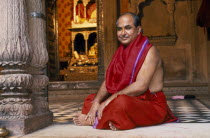 Hindu priest in red robes sitting cross legged on temple floor.