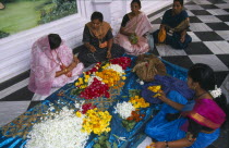 Women wearing colourful saris making flower garlands  sitting on black and white checked floor.
