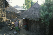 Young woman and children outside circular wooden house with thatched roof.