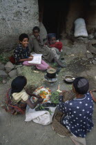 Group of woman and children sitting around cooking pot on stove outside building doorway.