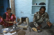 Woman cooking chapatis outside over simple  open top stove and serving them to a man sitting on the ground beside her.