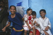 Four smiling children leaning against bicycle with blue wall behind.