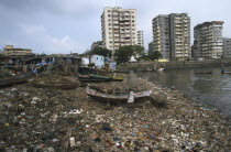 Polluted shoreline covered with rubbish with shanty housing and high-rise buildings behind.