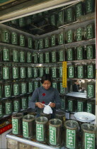 Young woman serving a customer partly seen reflected in mirrored wall behind her  in a shop selling Chinese medicines stored in jars on surrounding shelves.