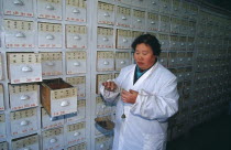 Woman weighing Chinese herbal medicines on hand held scales in front of wall of drawers.