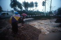 Torrent of flood water beneath bridge  watched by people with umbrellas.