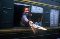 Old woman reaching out of the window of a train carriage with a straw hat in her outstretched hand.