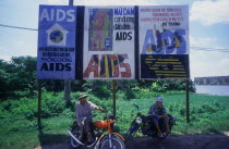 Two young men sitting on their motorcycles under a billboard of aids awareness posters.