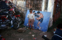 Small boy looking at painting of boxers propped against the wall in a motorcycle yard.