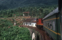 View from train along carriages as it crosses a bridge surrounded by lush vegetation