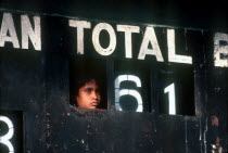 Young boy peering out through Cricket scoreboard to watch game