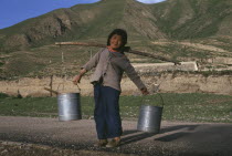 Child carrying water buckets on the ends of a stick over sholders.
