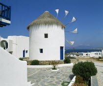 White windmill with thatched roof  sails have flags at tips  blue door  white town buildings & coast beyond. nr.06.053579.00