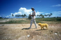 Man walking a dyed yellow dog near the beach with palms lining the horizon
