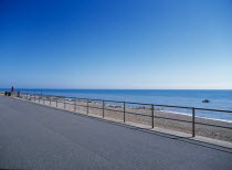 View from path across railings towards shingle beach and calm blue sea.