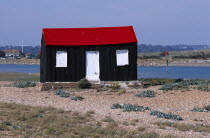 Rye harbour. Red and black corrugated hut with white doors built on the shingle with the harbour inlet behind.