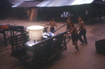A group of young boys pushing a handcart loaded with large containers in heavy rain.