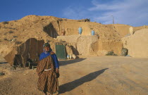 Portrait of a Berber woman standing outside her home which is dug out of a hillside