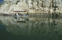 Painted tourist boat on the Narmada River passing sheer cliffs reflected in water.