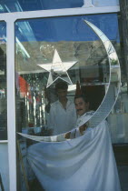 Barbers shop with star and moon of the Pakistan flag inlaid on the window in mirror glass reflecting the photographer.  Customer and barber seen inside